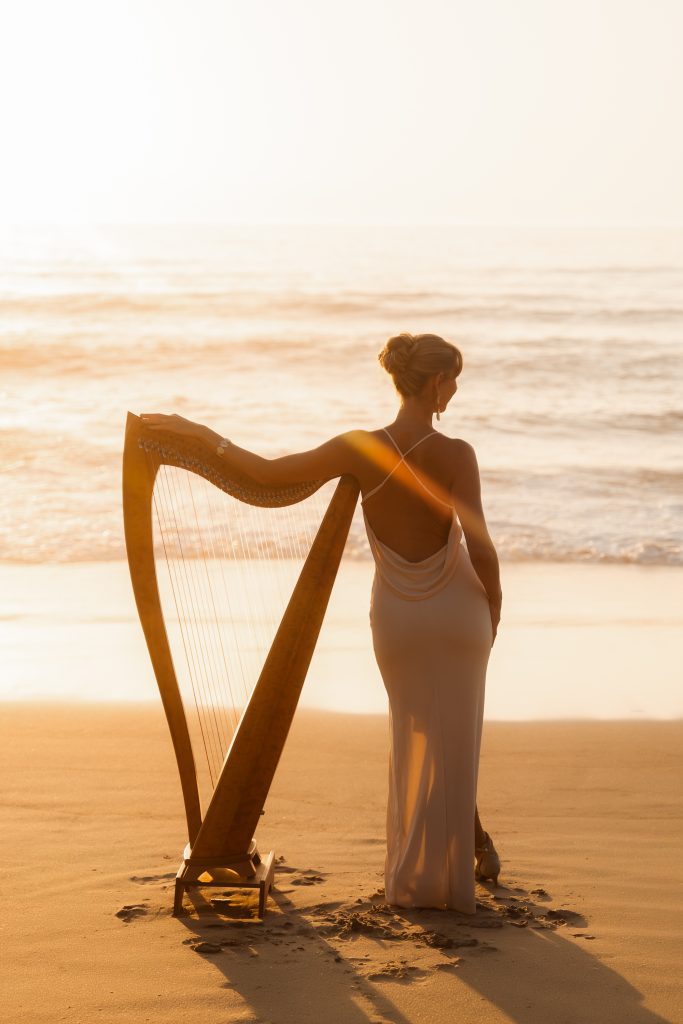 Danielle Riegel standing with her harp on the beach facing the ocean
